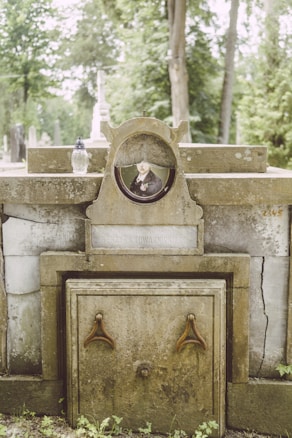 An old, weathered tombstone with a photograph of a man in formal attire is embedded in the center. The tombstone features inscriptions and has signs of aging, like moss and cracks. There is a small, decorative vase or candle holder placed on the stone. The background shows a lush, green cemetery with several trees and other gravestones.