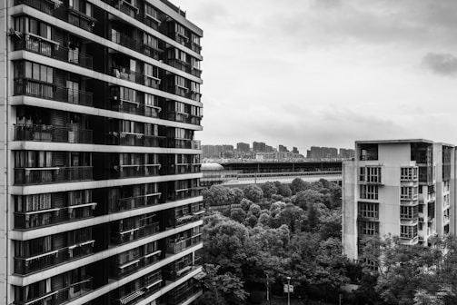 Two large residential buildings are prominently visible, with balconies and windows suggesting multiple apartments. In the background, a train or metro line runs parallel to the buildings, with a series of smaller city buildings under a cloudy sky. Dense greenery is also visible between and around the structures.