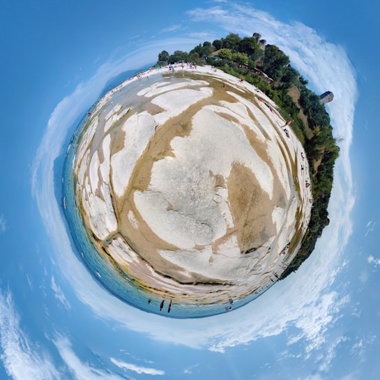 An artistic portrayal of a rocky beach and lush greenery captured with a fisheye lens, creating a planet-like effect. The sky is clear with a few clouds, and there are people scattered across the landscape enjoying the scenery.
