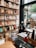A cozy bookstore interior featuring shelves filled with neatly arranged books and notebooks, a wooden stool, and a desk with assorted stationery items. A large window allows natural light to illuminate the space, offering a view of the street outside with visible trees and a parked vehicle.