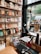 A cozy bookstore interior featuring shelves filled with neatly arranged books and notebooks, a wooden stool, and a desk with assorted stationery items. A large window allows natural light to illuminate the space, offering a view of the street outside with visible trees and a parked vehicle.