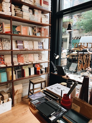 A cozy bookstore interior featuring shelves filled with neatly arranged books and notebooks, a wooden stool, and a desk with assorted stationery items. A large window allows natural light to illuminate the space, offering a view of the street outside with visible trees and a parked vehicle.