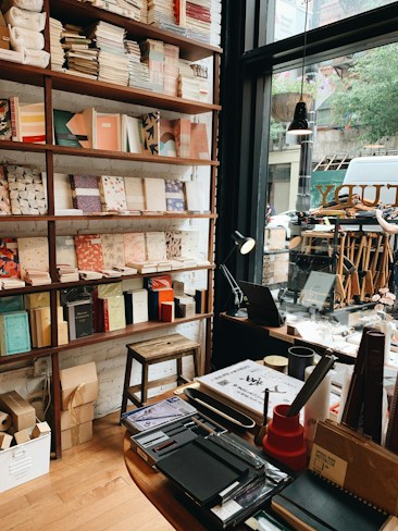 A cozy bookstore interior featuring shelves filled with neatly arranged books and notebooks, a wooden stool, and a desk with assorted stationery items. A large window allows natural light to illuminate the space, offering a view of the street outside with visible trees and a parked vehicle.