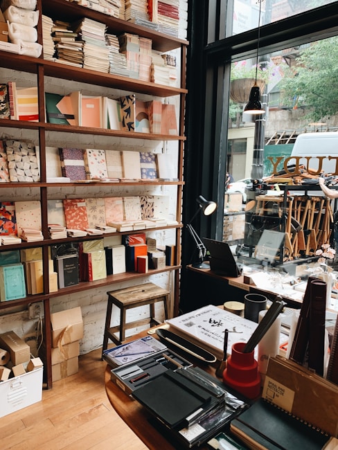 A cozy bookstore interior featuring shelves filled with neatly arranged books and notebooks, a wooden stool, and a desk with assorted stationery items. A large window allows natural light to illuminate the space, offering a view of the street outside with visible trees and a parked vehicle.