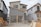 A partially constructed two-story house with a garage door frame is visible. The exterior features a combination of gray brick and unfinished walls covered with Tyvek wrap. To the side, there's construction machinery and building materials scattered on the ground.