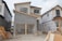 A partially constructed two-story house with a garage door frame is visible. The exterior features a combination of gray brick and unfinished walls covered with Tyvek wrap. To the side, there's construction machinery and building materials scattered on the ground.