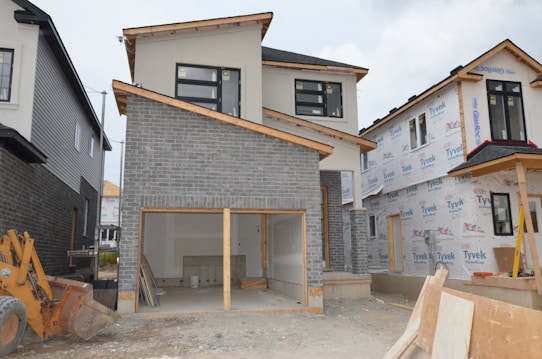A partially constructed two-story house with a garage door frame is visible. The exterior features a combination of gray brick and unfinished walls covered with Tyvek wrap. To the side, there's construction machinery and building materials scattered on the ground.