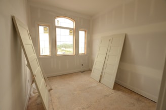 An empty room with unfinished drywall and three uninstalled white doors leaning against the walls. The room features three large windows with wooden frames and an arched central window, allowing natural light to flood in and revealing a view of the outside.