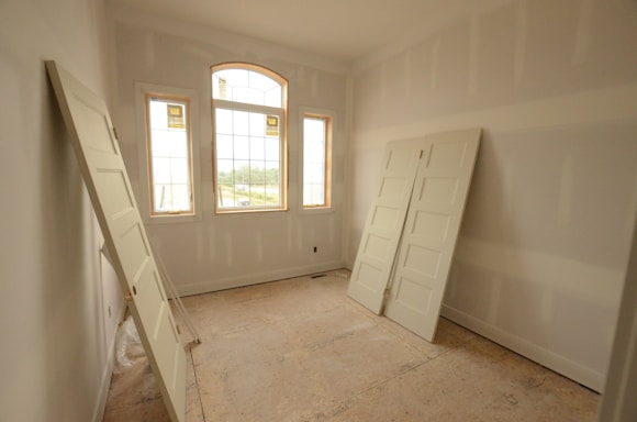An empty room with unfinished drywall and three uninstalled white doors leaning against the walls. The room features three large windows with wooden frames and an arched central window, allowing natural light to flood in and revealing a view of the outside.