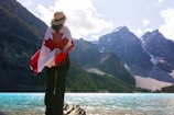 A person stands on a rock by a picturesque turquoise lake, draped in a Canadian flag, with tall, rocky mountains in the background under a partly cloudy sky. The individual is wearing a hat, with their back facing the camera, looking towards the mountains.