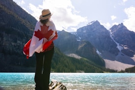 A person stands on a rock by a picturesque turquoise lake, draped in a Canadian flag, with tall, rocky mountains in the background under a partly cloudy sky. The individual is wearing a hat, with their back facing the camera, looking towards the mountains.