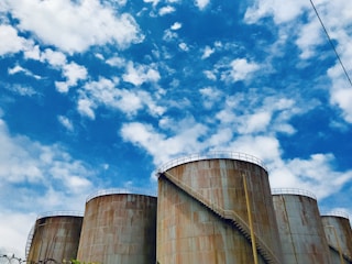 Several large industrial storage tanks are seen against a backdrop of a bright blue sky with scattered clouds. The tanks have a rusted appearance and feature metal staircases running along their sides.