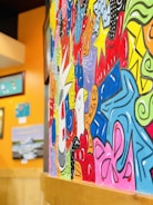 A vibrant and colorful mural featuring a mix of abstract designs and patterns, with swirling shapes and bold outlines against a backdrop of vivid colors like red, blue, yellow, and green. Various framed pieces of art can be seen hung on a bright orange wall nearby.