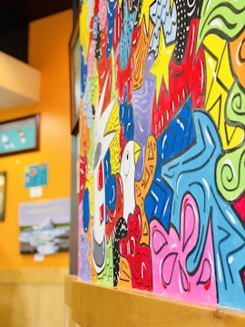 A vibrant and colorful mural featuring a mix of abstract designs and patterns, with swirling shapes and bold outlines against a backdrop of vivid colors like red, blue, yellow, and green. Various framed pieces of art can be seen hung on a bright orange wall nearby.