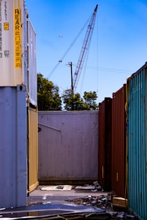 A construction crane towers above a row of shipping containers lined up closely together. Various colors of the containers, including yellow, red, and teal, contrast with the clear blue sky. There is some debris and litter like cardboard on the ground between the containers, and a power line runs across the scene. Trees are visible in the background, and a bird is flying high in the sky near the crane.