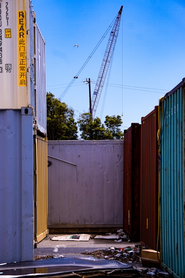 A construction crane towers above a row of shipping containers lined up closely together. Various colors of the containers, including yellow, red, and teal, contrast with the clear blue sky. There is some debris and litter like cardboard on the ground between the containers, and a power line runs across the scene. Trees are visible in the background, and a bird is flying high in the sky near the crane.