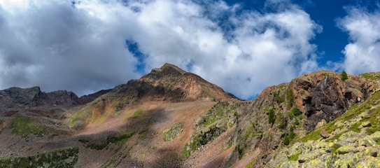 A rugged mountain landscape with a prominent peak under a partly cloudy sky. The terrain is rocky with patches of greenery and varying shades of earth tones, creating a dramatic contrast with the blue and white sky.