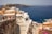 A scenic seaside village with whitewashed buildings and a deep blue sea in the background. Tourists walk along narrow pathways lined with colorful flowers and quaint shops. The landscape includes rugged cliffs and architectural elements typical of a Mediterranean setting.