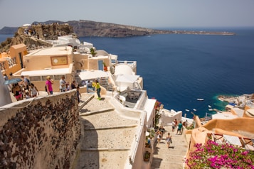 A scenic seaside village with whitewashed buildings and a deep blue sea in the background. Tourists walk along narrow pathways lined with colorful flowers and quaint shops. The landscape includes rugged cliffs and architectural elements typical of a Mediterranean setting.