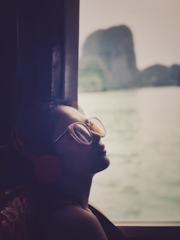 A person is sitting indoors near a window, wearing large sunglasses and headphones, and appears to be relaxed. The window provides a view of a distant rock formation and a body of water, suggesting a serene outdoor landscape.