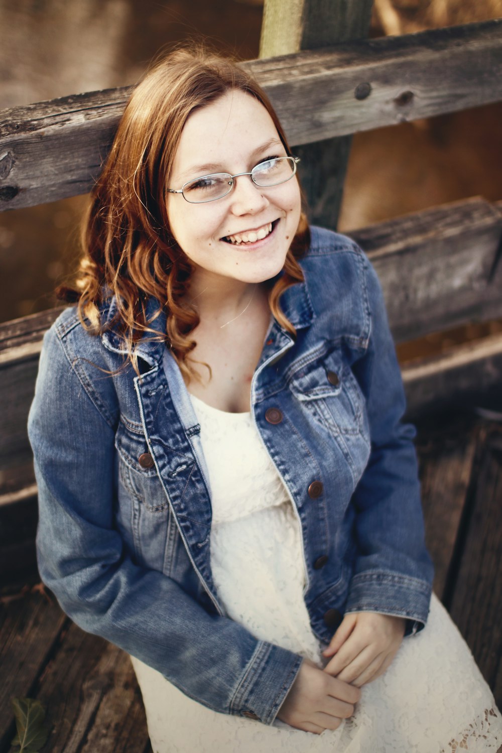 mulher sorridente vestindo jaqueta jeans azul e óculos sentada no banco de madeira marrom