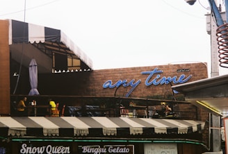 A café with an outdoor seating area under a striped awning, featuring a brick wall with a neon sign saying 'any time'. People are sitting at tables on the patio, creating a casual, laid-back atmosphere.