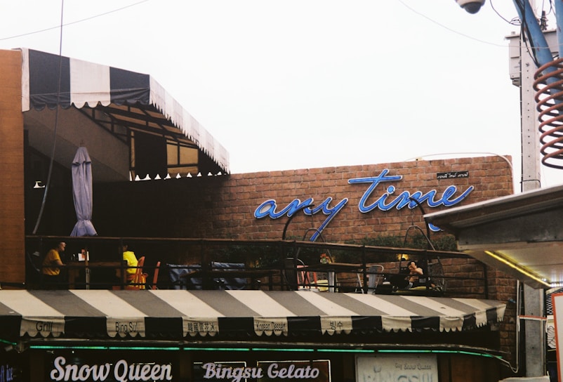 A café with an outdoor seating area under a striped awning, featuring a brick wall with a neon sign saying 'any time'. People are sitting at tables on the patio, creating a casual, laid-back atmosphere.