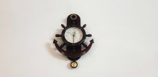 A wall clock designed to resemble a ship's helm and anchor. The clock is mounted on a white wall and features Roman numerals on its face. The helm's wheel spokes are visible surrounding the clock, and an anchor shape extends below it.