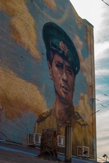 A large mural of a person wearing a military uniform is painted on the side of a brick building. The person is depicted with a serious expression, wearing a dark green hat adorned with a badge. The background features clouds painted in shades of beige and blue, blending with the aged texture of the bricks.