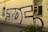 A large piece of graffiti with the letters 'TDF' in bold, outlined with a dark purple color, adorns the side of an old beige building. The building has a worn appearance, with a damaged window featuring broken panes and green weeds growing at the base of the wall.
