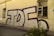 A large piece of graffiti with the letters 'TDF' in bold, outlined with a dark purple color, adorns the side of an old beige building. The building has a worn appearance, with a damaged window featuring broken panes and green weeds growing at the base of the wall.