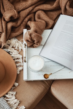 A cozy setup featuring a cream-colored sofa adorned with a brown knit blanket and a fringed throw. An open book, a mug with frothy contents, and a spoon are neatly arranged on a white tray. A wide-brimmed hat is placed on the sofa.