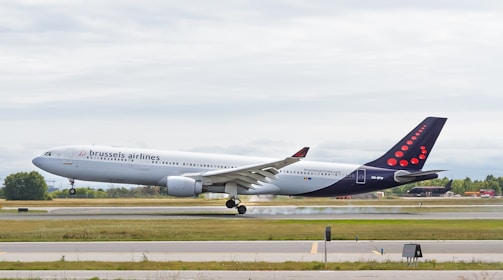A commercial airplane from Brussels Airlines is landing on a runway, with its landing gear deployed. The aircraft is touching down, and there is a visible puff of smoke from the tires hitting the ground. The background features a cloudy sky and some greenery surrounding the airfield.