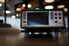 A Keithley data acquisition and multimeter system placed on a table. The device features a large screen with surrounding control buttons and various input ports. The background is softly blurred, suggesting an office or laboratory environment, with circular lights visible.