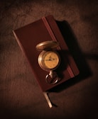 An antique gold pocket watch rests atop a closed brown leather-bound notebook. The watch's cover is open, revealing its vintage face with Roman numerals. Both items are set upon a textured dark brown fabric, creating a warm and nostalgic atmosphere.