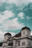 A colonial-style building with prominent domes and intricate architectural details. The building's facade is white with decorative elements, and the sign reads 'Bank Indonesia'. The sky above is filled with scattered clouds and has a teal hue.