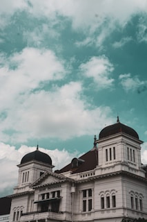 A colonial-style building with prominent domes and intricate architectural details. The building's facade is white with decorative elements, and the sign reads 'Bank Indonesia'. The sky above is filled with scattered clouds and has a teal hue.