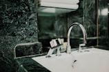 A modern bathroom sink features a shiny chrome faucet set against a background of dark green marble tiles. Above the sink, a glass shelf holds neatly stacked white towels.