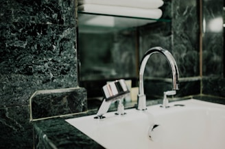 A modern bathroom sink features a shiny chrome faucet set against a background of dark green marble tiles. Above the sink, a glass shelf holds neatly stacked white towels.