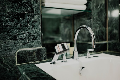 A modern bathroom sink features a shiny chrome faucet set against a background of dark green marble tiles. Above the sink, a glass shelf holds neatly stacked white towels.