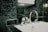 A modern bathroom sink features a shiny chrome faucet set against a background of dark green marble tiles. Above the sink, a glass shelf holds neatly stacked white towels.
