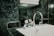 A modern bathroom sink features a shiny chrome faucet set against a background of dark green marble tiles. Above the sink, a glass shelf holds neatly stacked white towels.