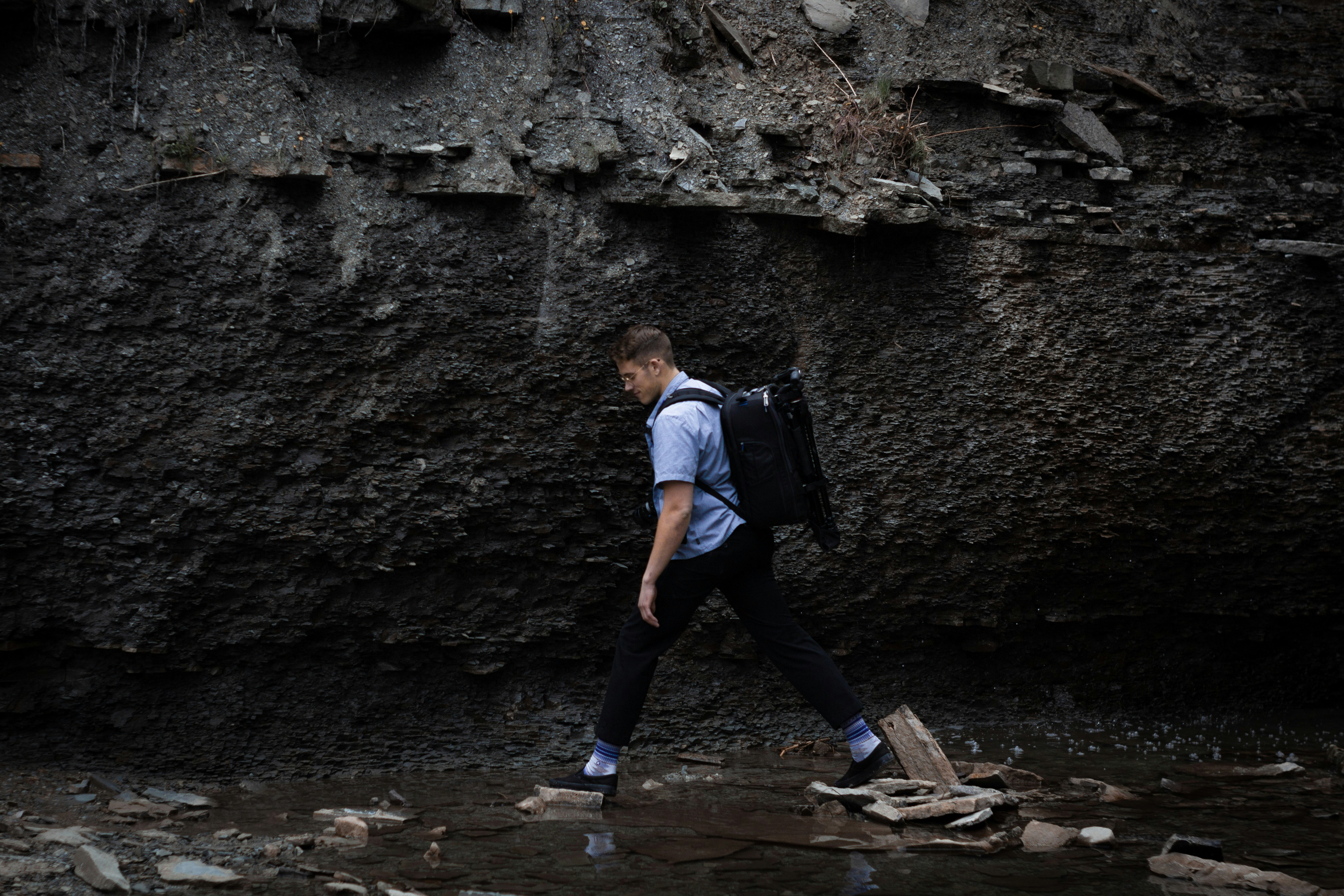 man with black backpack stepping on rocks near river