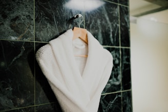 A white bathrobe is neatly hung on a wooden hanger against a textured dark green marble tiled wall. The robe appears plush and soft, suggesting comfort and luxury.
