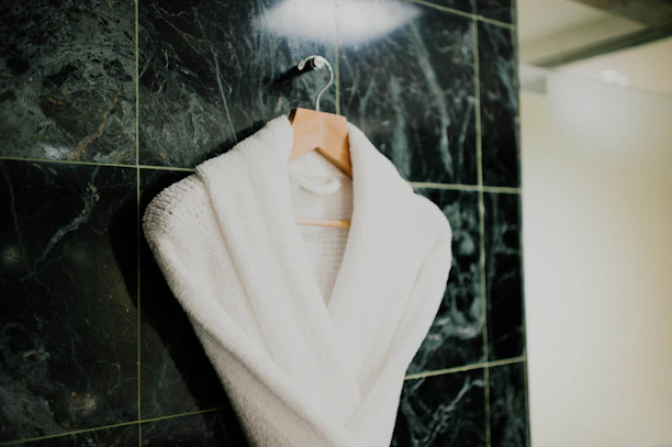 A white bathrobe is neatly hung on a wooden hanger against a textured dark green marble tiled wall. The robe appears plush and soft, suggesting comfort and luxury.