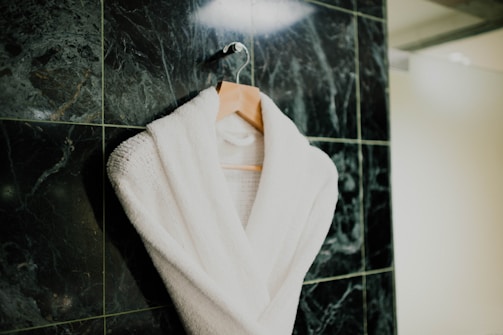A white bathrobe is neatly hung on a wooden hanger against a textured dark green marble tiled wall. The robe appears plush and soft, suggesting comfort and luxury.