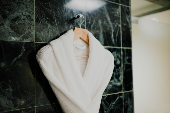 A white bathrobe is neatly hung on a wooden hanger against a textured dark green marble tiled wall. The robe appears plush and soft, suggesting comfort and luxury.