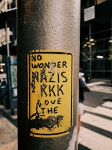 A yellow sticker on a weathered metal pole featuring bold black text that appears political in nature. The background shows an urban environment with blurred buildings and a street.