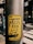 A yellow sticker on a weathered metal pole featuring bold black text that appears political in nature. The background shows an urban environment with blurred buildings and a street.