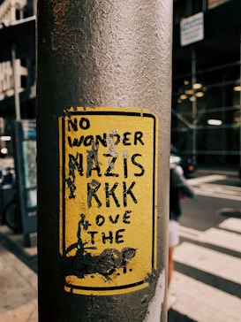 A yellow sticker on a weathered metal pole featuring bold black text that appears political in nature. The background shows an urban environment with blurred buildings and a street.